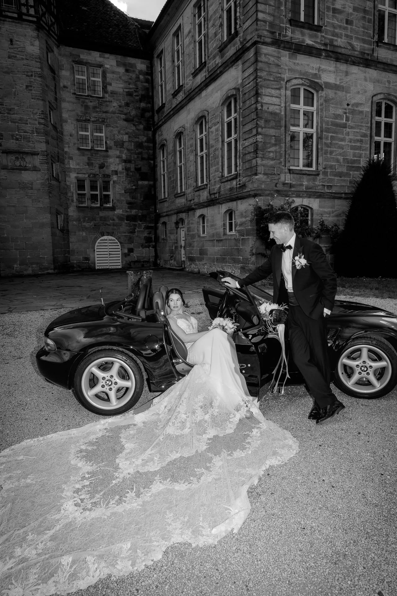 Glamorous bride and groom at convertible in front of Schloss Eyrichshof in black and white
