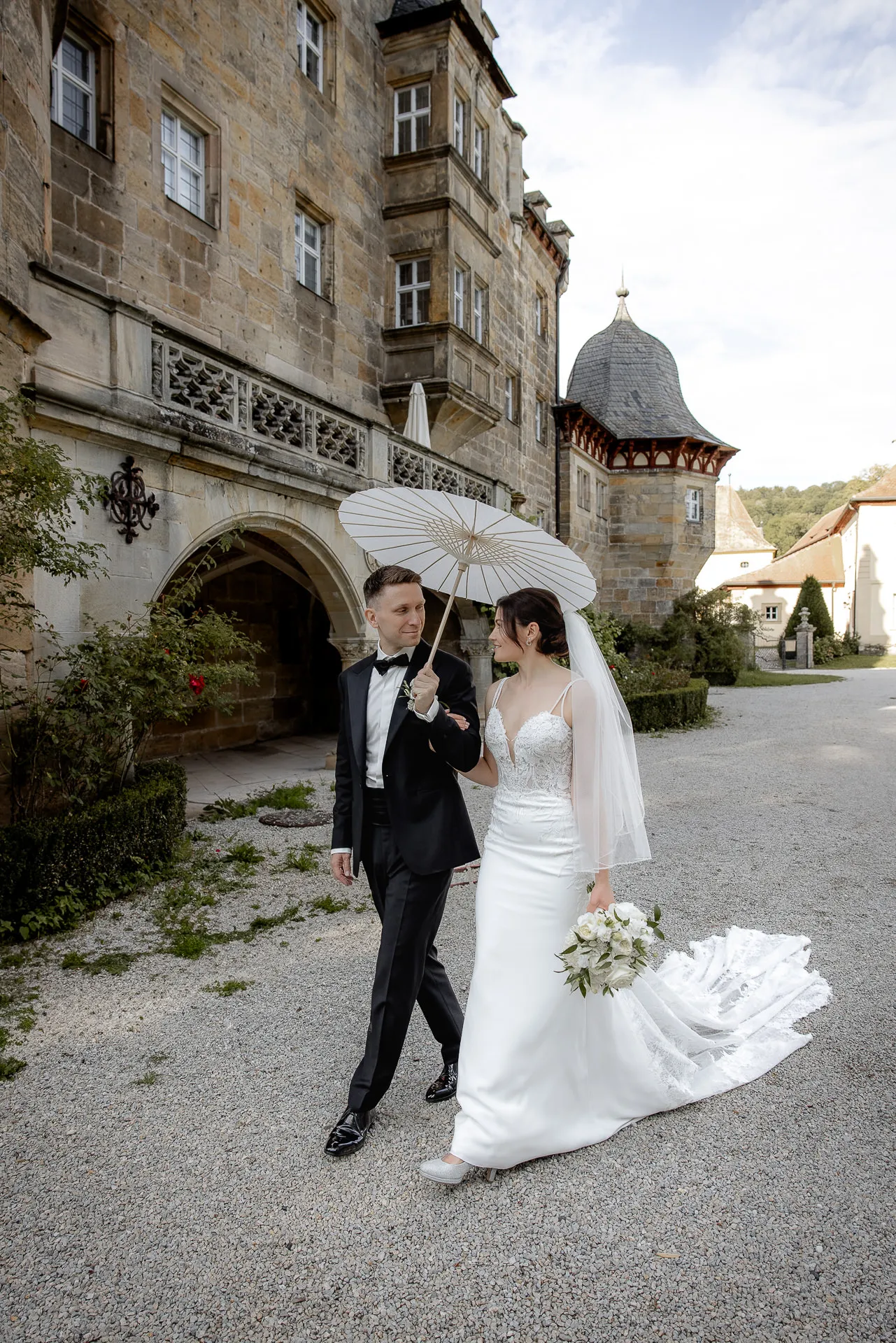 Bride and groom with white parasol walking through Schloss Eyrichshof courtyard at wedding