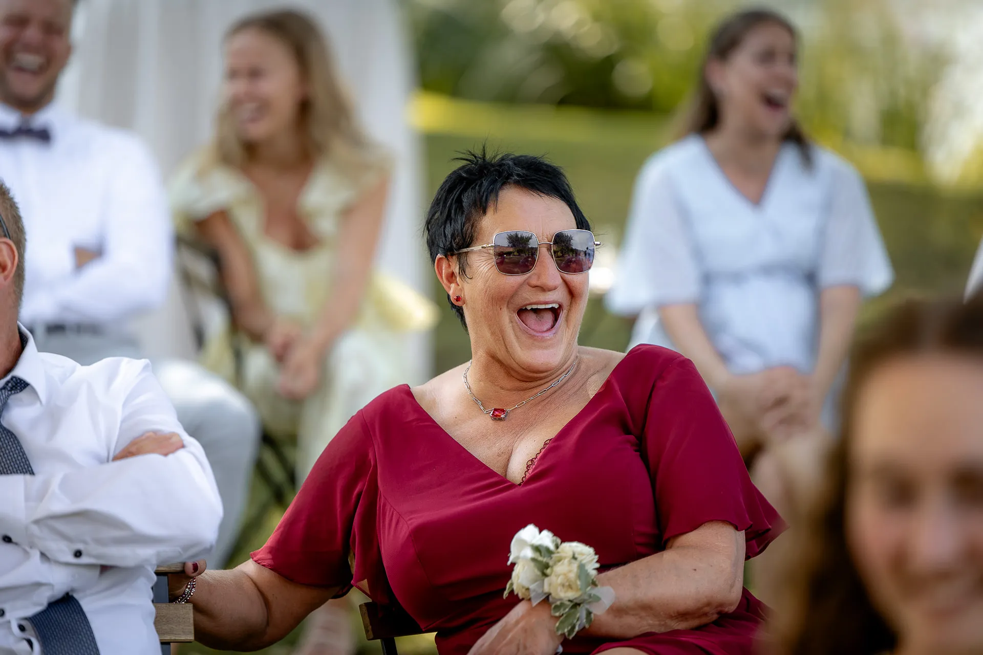Laughing wedding guest in burgundy dress with sunglasses holding bouquet