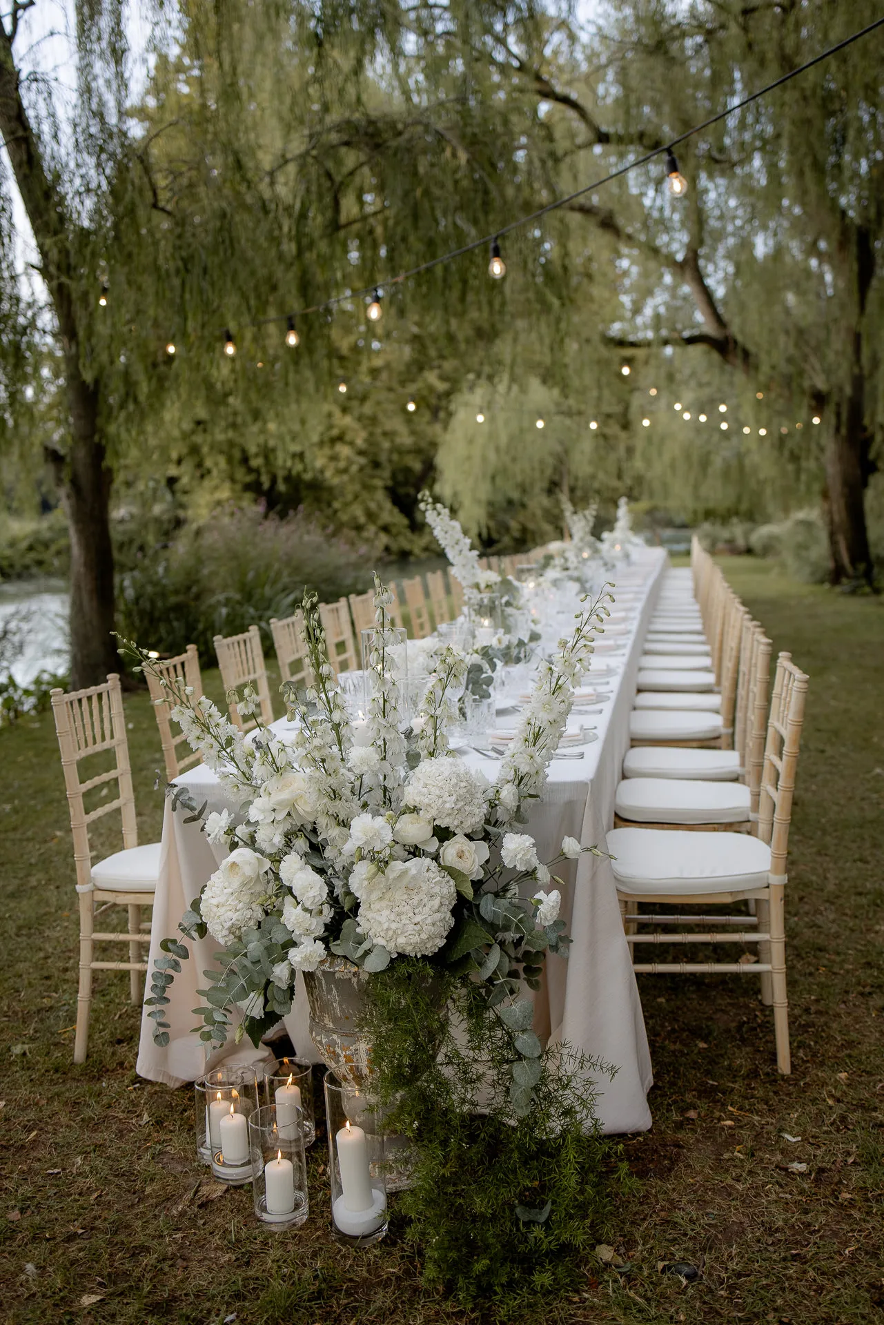 Romantic evening wedding table with white floral decor and lights under willows