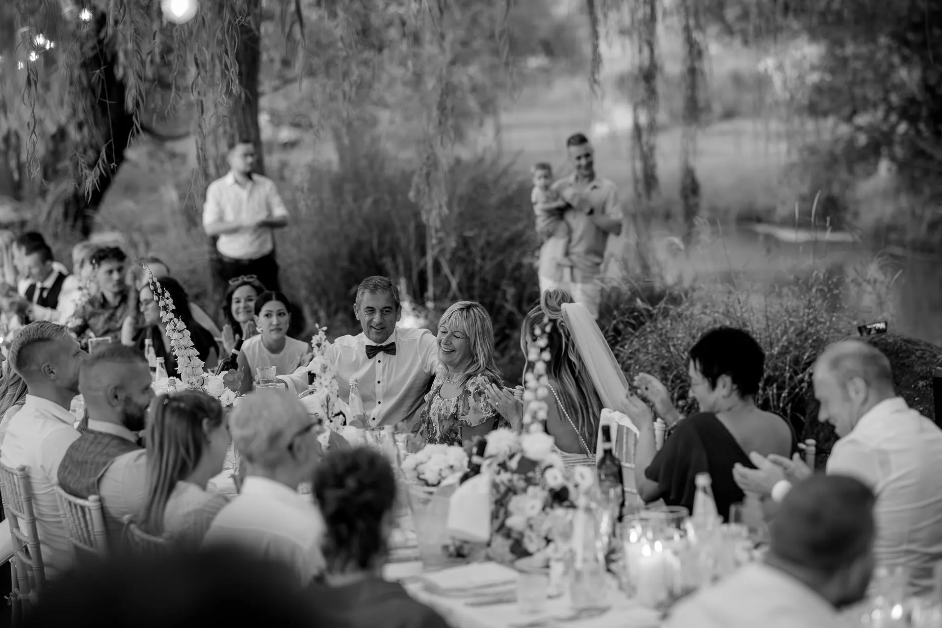 Wedding party celebrating at long table under willows by riverside at twilight