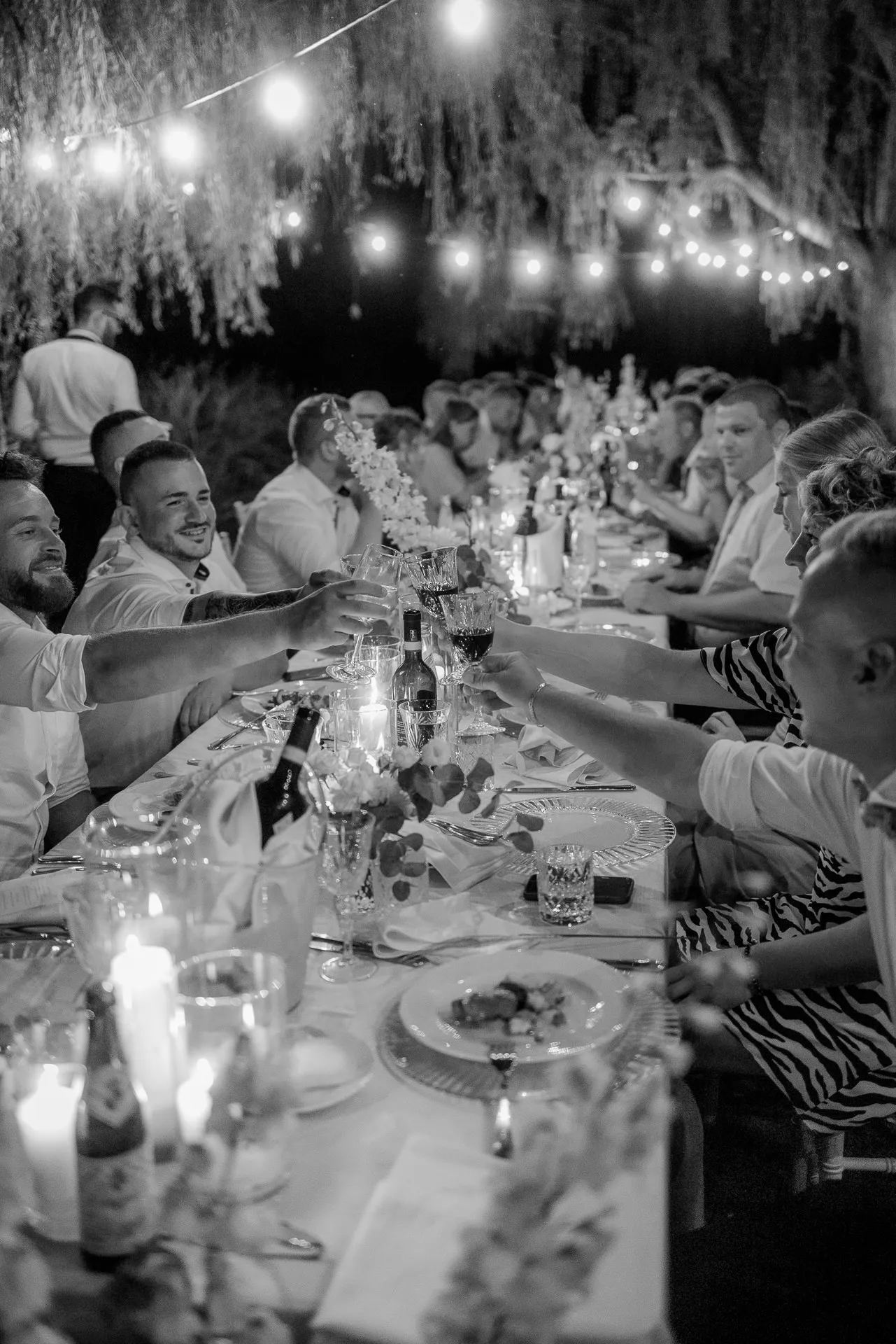 Wedding guests cheerfully toasting at evening dinner under lights and willows