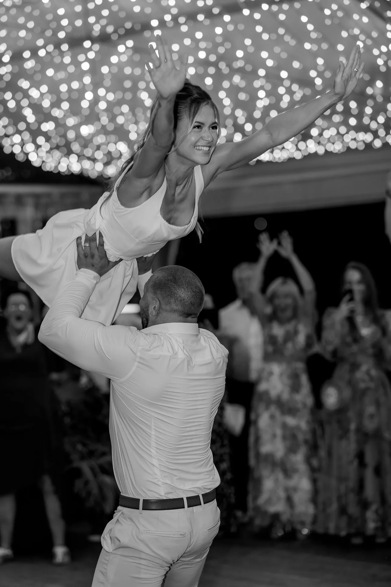 Groom lifts jubilant bride in spectacular dance before string lights background