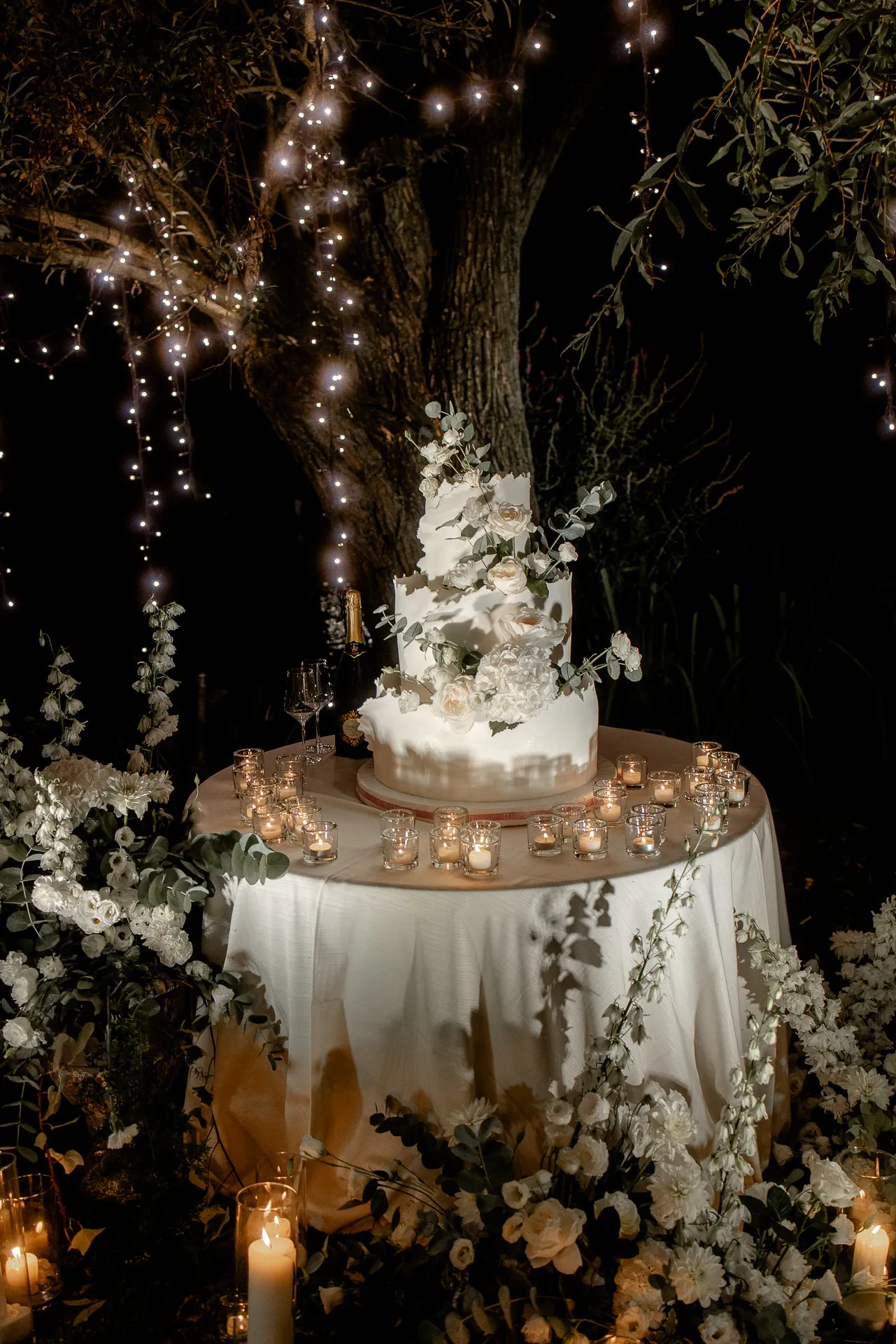 Three-tier wedding cake with eucalyptus decor surrounded by candles and flowers