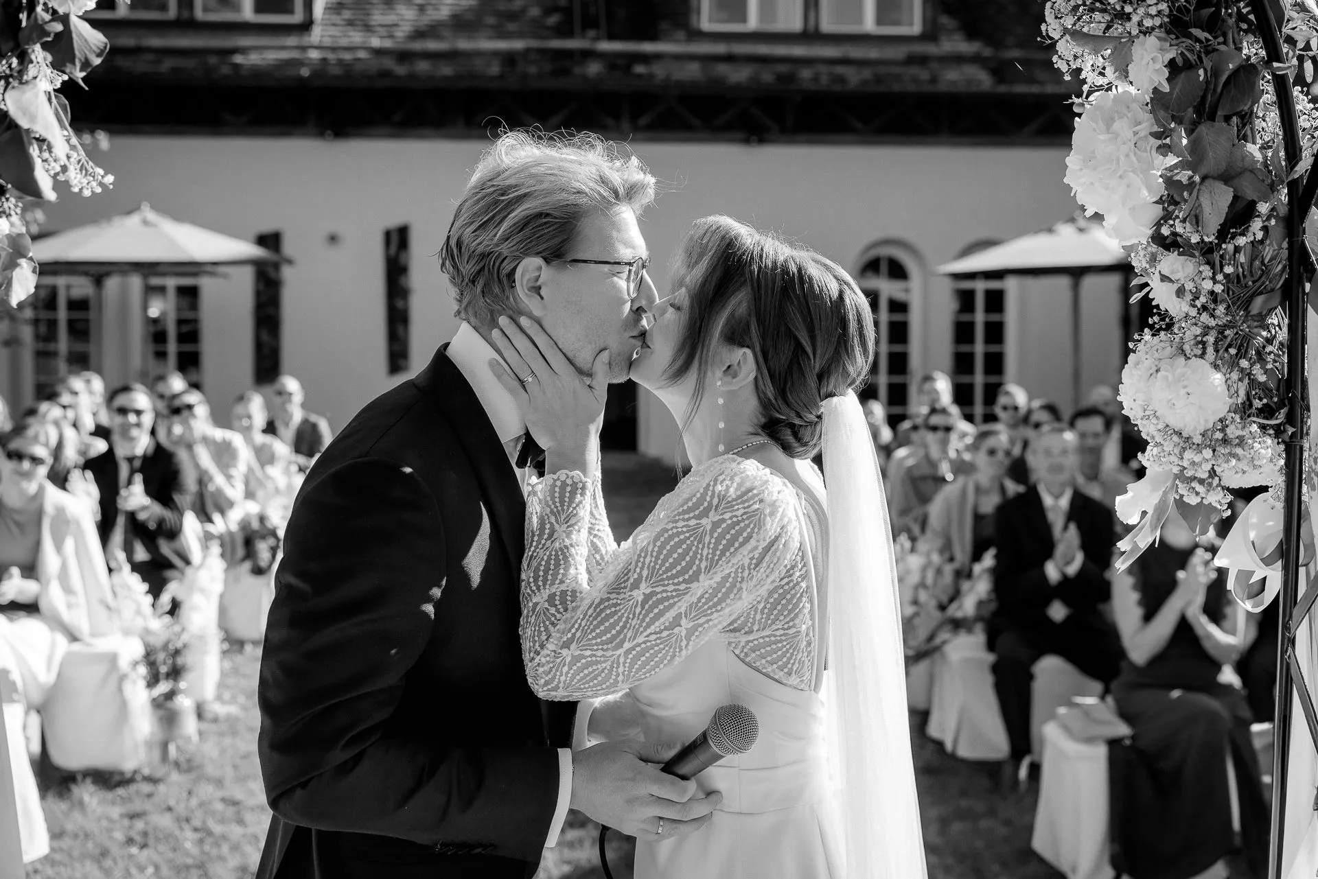 Bride and groom kissing after ceremony with applauding guests in black and white